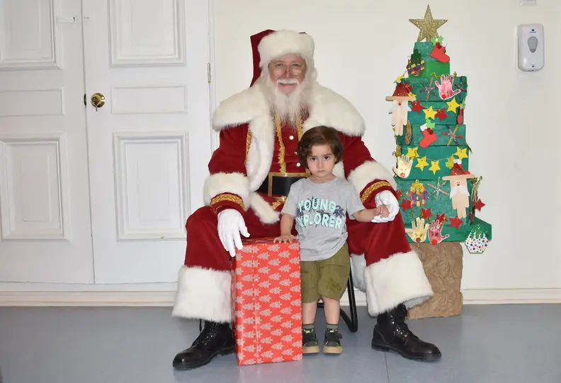 Niño tomándose una foto con el viejito pascuero, y con su regalo, de fondo un árbol de navidad, en una sala blanca